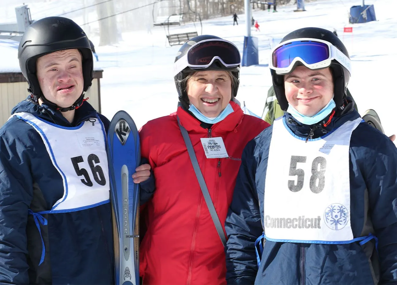 Two athletes standing with a partner outside in the snow wearing skiing attire.
